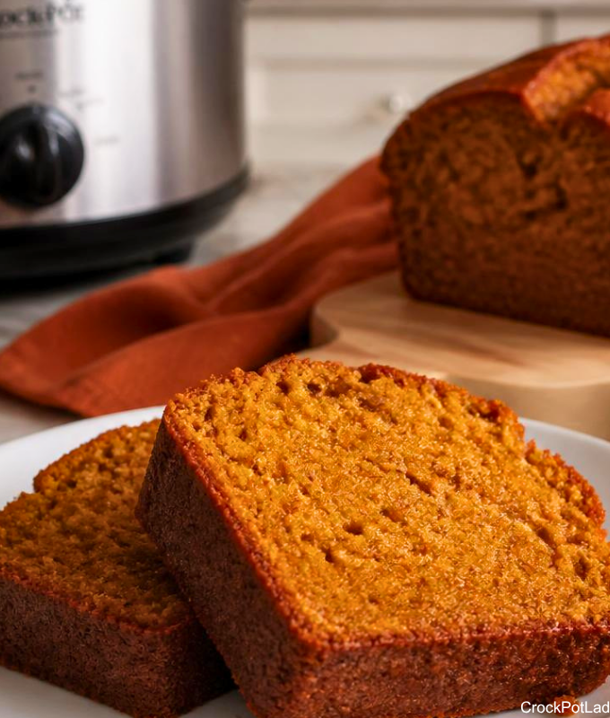 Two close-up slices of moist Crock-Pot pumpkin bread on a white plate, with a whole loaf and a slow cooker in the soft-focus background.